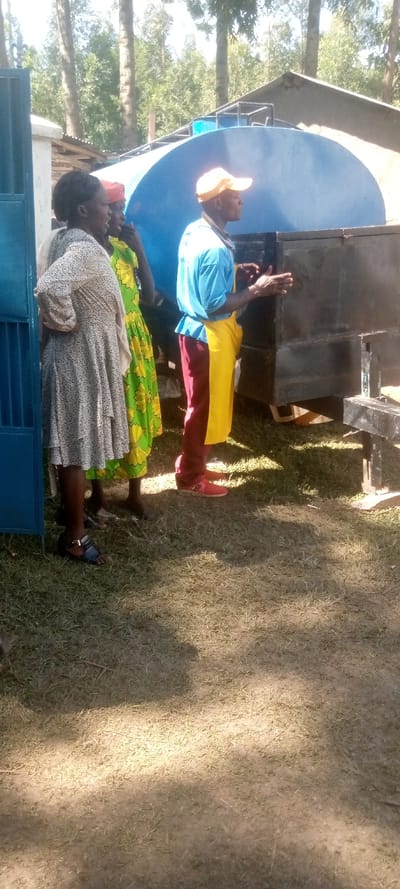 The image shows three people standing in front of a blue water tank on a dirt lot in Kivaywa, Kenya. The location is confirmed using the provided latitude and longitude coordinates.  The people are all dressed in casual clothing. The person on the left is wearing a white dress, the person in the middle is wearing a green and yellow dress, and the person on the right is wearing a blue shirt, yellow apron, and maroon pants. The man on the right is pointing his right hand at something behind the camera. The ground is covered in dry grass and dirt. There is a blue door in the background on the left side of the image. There is a large blue water tank in the background that is on a metal frame. The tank is partially covered by a metal roof. There is a building in the background on the right side of the image.  The scene is bright and sunny. The sky is clear. There are trees in the background.
