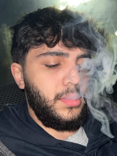 A male individual is depicted in a close-up, chest-up shot, located in Al-Muhajerin Municipality, Syria. The subject has dark, curly hair and a full, dark beard. His eyes are closed. A plume of white-grey smoke is visibly exhaling from his mouth, partially obscuring the right side of his face and circulating around his head. He is wearing a dark, possibly navy, hooded sweatshirt over a lighter grey garment. The background is dark and out of focus, revealing a dark, textured surface to the left, consistent with an interior or vehicle environment.
