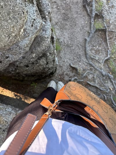 The image shows a close-up perspective of a person sitting outdoors, likely on a rocky terrain with exposed tree roots. The person is wearing dark pants and white shoes. They are holding a brown and tan Guess handbag, which has a distinctive "G" logo pattern. The handbag's zipper is partially open, revealing the interior. The background consists of large grey rocks and the ground is covered with dirt, small stones, and patches of green grass. The lighting suggests it is daytime, with sunlight casting shadows. The specific location in Szklarska Poręba, Poland, is not identifiable from the visual elements alone.