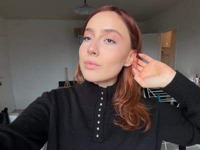 This indoor photograph depicts a young adult female, positioned from the chest up and slightly off-center to the right, within what appears to be a residential room in Reims, France. She has long, wavy, reddish-brown hair and is wearing a black, ribbed knit sweater with a high collar featuring a partial button-up placket fastened with five small, silver-colored spherical buttons. Her visible right hand, with light-colored manicured nails, is raised, touching her hair near her right earlobe, which features a small silver-colored stud earring. Her face exhibits light makeup, including pink eyeshadow and lip gloss. The background consists of plain, light grey walls. An overhead grey metal dome-shaped pendant lamp is visible in the upper center. To the left, a partial white object and a dark object are visible. In the mid-ground right, a wooden tripod-like stand and a black rectangular object are present. Further right, a white doorframe leads into another room, revealing a white wall and a white appliance. A white metal drying rack and several small containers on a surface are partially visible on the far right.