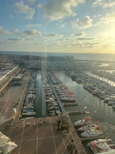 This high-angle shot captures a bustling marina in Herzliya, Israel, under a bright, partly cloudy sky. The image appears to be taken from a tall building or elevated viewpoint, looking down upon the extensive harbor and coastline.
The sky, occupying a significant portion of the image, is a pale blue with scattered white cumulus clouds. A strong glare from the sun is visible in the upper left, indicating a bright, sunny day, likely mid-morning or afternoon.
The central feature is the marina, densely packed with numerous boats, predominantly sailboats, docked in neat rows. Their tall masts create a forest-like appearance against the water. Many boats have their sails furled or covered. The calm water reflects light and the hulls of the vessels. Visible text on some boat hulls includes "Sea-Gal" and partially "Racing". Several Israeli flags are seen flying from the masts of some boats, a clear indicator of the location.
Adjacent to the water is a wide, paved pier or boardwalk, featuring a distinctive brick pattern. Small figures of people can be seen walking along this promenade, adding a sense of scale and activity. Along the landward side of the marina, modern buildings with intricate, geometric architectural designs line the coast. On the pier, there are various temporary structures, including white tent-like covers, as well as scaffolding or ladders, suggesting ongoing work or events. Street furniture, such as signposts and bins, is also scattered across the paved area.
The overall scene conveys a vibrant and active maritime environment, characteristic of a popular coastal destination.