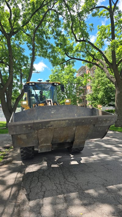 A large, heavy-duty yellow construction vehicle, specifically a backhoe loader, is parked on an asphalt street in Oakville, Canada. The vehicle's front loader bucket is positioned forward and slightly lowered. Inside the cab, a person wearing a yellow reflective vest and a dark shirt is visible, appearing to be the operator. The scene is set on a bright, sunny day with dappled sunlight filtering through the dense green canopy of mature trees that line the street. In the background, a multi-story brick apartment building with white balconies is partially visible amidst the foliage. The street surface shows signs of wear and cracking.