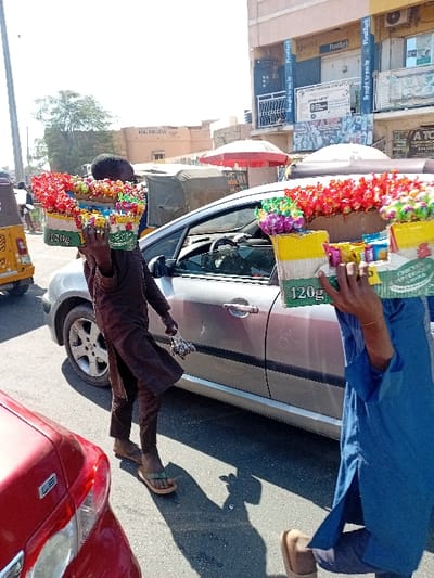 A daylight urban street scene in Kabuga, Nigeria, depicts two male street vendors navigating a paved road. Each individual carries a rectangular tray loaded with brightly colored, individually packaged snacks or candies balanced on their head. The vendor on the left, partially obscured, wears a dark long-sleeved garment, dark trousers, and light green flip-flops, while holding an unidentifiable bundled object in his left hand. The vendor on the right, also partially obscured, wears a blue long-sleeved tunic, light trousers, and light brown slip-on shoes. A silver sedan is positioned between the two vendors, with a red vehicle partially visible in the extreme foreground-left. Strong shadows are cast on the asphalt, indicating bright sunlight. The background features commercial buildings, including a multi-story structure prominently displaying "FirstBank" branding in blue and yellow, along with various advertisements and a red parasol. A yellow tricycle is partially visible on the far left.