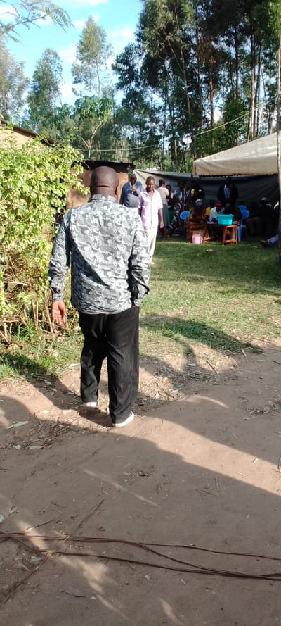 The scene is a dirt road in Kipkarren, Kenya (latitude: 0.599, longitude: 34.957). A man wearing a grey and black shirt, black pants, and black shoes is walking away from the camera on a dirt road. He is walking in a field with grass. Behind him are several people sitting in chairs under a white tent. The area appears to be rural, with trees and shrubs in the background. There is a dirt path leading to a small, one-story building in the background. The man is the only one in focus. There are several other people out of focus in the background. There is a shadow on the dirt road behind the man.