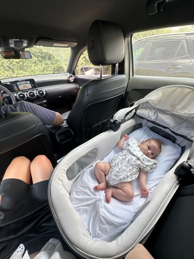 The image depicts the interior of a car, likely in Carini, Italy, with a baby lying in a bassinet. The baby is wearing a white romper with a floral pattern and a bib with hearts. A person's legs, clad in a black skirt and white lace, are visible in the foreground. Another person's arm and leg are visible in the middle ground, suggesting they are driving the car. The car's dashboard, steering wheel, and infotainment system are partially visible. Outside the car, lush green foliage can be seen through the windows. A dark-colored SUV is parked beside the car. The overall scene suggests a family outing or travel.
