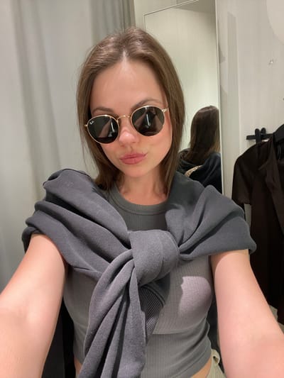 A young woman, with brown hair and wearing gold-framed Ray-Ban sunglasses, is depicted from the upper chest in a modern indoor setting, specifically a fitting room, located in the city of Pompei, Italy. Her lips are slightly puckered. She is dressed in a light grey ribbed tank top, and a dark grey sweatshirt is tied around her shoulders with the sleeves knotted at the front. Her bare arms are visible. A mirror in the background reflects the back of her head and shoulders, as well as a dark garment hanging on a clothing rack to the right. The environment features plain, light-colored walls and uniform artificial lighting.