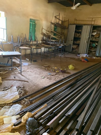 The image shows the interior of a room in Khartoum, Sudan. The room is in disarray with numerous overturned chairs and desks. There is a large metal shelving unit on the right side of the image, mostly filled with files. Several stacks of metal bars are scattered across the floor. Some paper and a plastic bag are on the floor in the lower left corner of the image. There is a ceiling fan in the corner of the room, with two windows behind the overturned desks, and a large window in the back left corner of the room. The walls of the room are painted a light yellow, with a light brown or beige floor. There are no people present in the image.  The location of the image is a room at the coordinates 15.552 latitude and 32.57 longitude in Khartoum, Sudan.