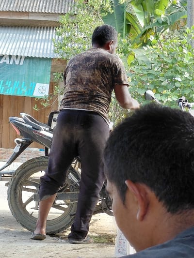 Two men are fixing a motorcycle. One man is wearing a patterned brown shirt and dark pants, while the other is wearing a blue shirt, his head bent over the motorcycle. A motorcycle tire is visible along with parts of its frame. A corrugated metal building and a green sign are in the background, along with trees. The scene is brightly lit.
