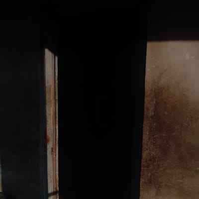 The image depicts a dimly lit scene with strong shadows and highlights, suggesting it was taken at either sunrise or sunset. A vertical element, possibly a door frame or pillar, is visible on the left, with a rough, weathered texture and hints of rust or discoloration. This element is partially illuminated by a shaft of light, casting a sharp shadow. To the right of this element is a large area of deep shadow, obscuring any details within it. Further to the right, a textured wall surface is visible, appearing somewhat dirty or weathered, with lighter specks and darker, irregular patches, possibly stains or discoloration. The overall environment appears to be outdoors, in an urban setting, and given the context provided, this scene is located in Ogbomosho, Nigeria. There are no people or discernible actions visible in the image.