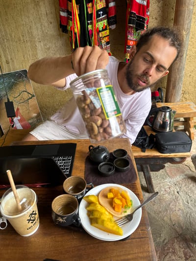 A man with brown hair and a beard is seated at a wooden table in Lũng Cú Commune, Vietnam. He is wearing a light pink t-shirt and white shorts. He is holding a clear jar filled with nuts.
On the table are:
* A laptop computer (black)
* A small teapot with four cups
* A white plate with a pancake topped with diced mango and slices of starfruit, along with a fork
* Two empty cups
* A paper cup with a clear lid and a straw.
In the background are:
* A shelf with colorful textiles
* A metal teapot and a black bag hanging on a wooden structure.
The walls are textured, possibly stone or stucco. A power adapter is visible below the table.