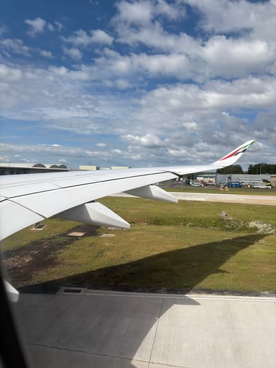 The image shows a view from inside an airplane, looking out over the wing and the airport grounds. The wing, painted white, takes up a large portion of the frame, and the Emirates logo is visible on the wingtip. The airport grounds below consist of a concrete surface and grassy areas. In the background, there are airport buildings and vehicles. The sky is visible with white clouds against a blue background. Based on the Emirates logo and the general appearance of the airport, the location is likely near Gatwick Airport, in the city of Crawley, United Kingdom.