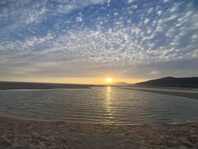 The image captures a tranquil beach scene at sunset in Tarifa, Spain. The sky is a canvas of blue, adorned with scattered, puffy white clouds, creating a textured and dynamic ceiling. The sun, a bright orb, is low on the horizon, casting a warm, golden glow that reflects off the calm waters of a shallow lagoon or river mouth. The water's surface is rippled, showing the gentle movement of the tide. In the foreground, the sandy beach exhibits the subtle undulations of its surface. Beyond the water, a distant hilly landscape is silhouetted against the colorful sky. On the crest of the hill, several wind turbines stand as silent sentinels, a characteristic feature of the Tarifa region. The overall impression is one of peaceful natural beauty at the end of the day.