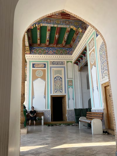 A person sits on a bench in a courtyard with ornate, colorful ceilings and walls. The architecture features intricate geometric patterns and Arabic calligraphy, indicative of Central Asian Islamic art. The ceiling is elaborately decorated with green, red, blue, and gold colors, featuring carved wooden beams. The walls are adorned with colorful tiled panels, some containing calligraphy. Sunlight streams into the courtyard, casting shadows on the tiled floor, suggesting it's daytime. The person, wearing sunglasses and a black outfit, appears relaxed. The setting looks like a historical site or a mosque courtyard in Samarkand, Uzbekistan, possibly a tourist area given the bench and the presence of shoes lined up near an entrance. No discernible weather conditions are apparent, but the light indicates a sunny day.