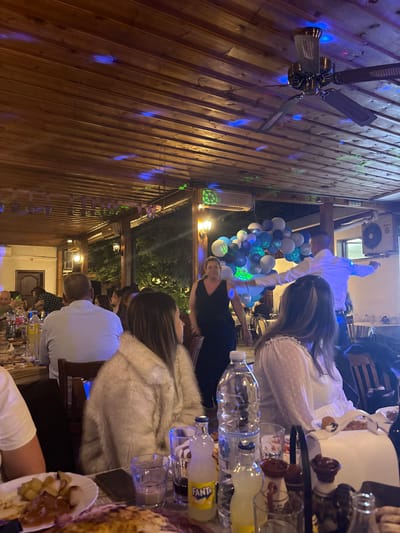 The image depicts an indoor gathering, possibly a celebration, at a restaurant in Karlovo, Bulgaria. The scene is dimly lit, with a wooden ceiling and exposed wooden beams above. Several people are seated at tables, engaged in conversation and dining. In the background, a man in a white shirt is dancing, with a backdrop of blue and white balloons. A woman in a black dress stands to his left, observing. There are bottles of beverages on the tables, along with plates of food. A ceiling fan is visible, and colored lights, primarily blue, are projected onto the ceiling, creating a festive atmosphere. The overall impression is one of a lively social event.