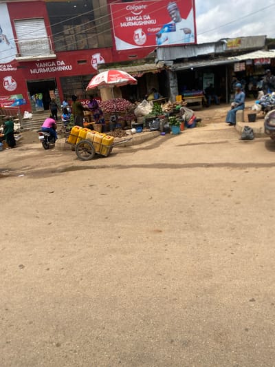 In Kabong, Nigeria, a busy street scene unfolds. A red building with a prominent "YANGA DA MURMUSHINKU" sign and Colgate advertisements stands as the backdrop. In front of the building, a market is in full swing. Several people are engaged in various activities: a person on a motorcycle wearing a pink shirt, vendors arranging produce, and customers browsing. Yellow containers are stacked on a cart, suggesting the transport of goods. The ground is a dirt road, indicative of a local marketplace. The environment is characterized by an outdoor market setting with stalls, goods, and a mix of people.