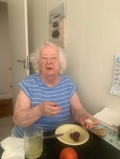 An elderly woman with white hair is sitting at a table, eating. She is wearing a blue and white striped shirt. In her right hand, she is holding a piece of bread with cheese. In front of her is a bowl with cherries, a peach, and a glass of lemonade. A calendar is visible on the wall behind her, with the dates showing "23", "24", "25", "26", "27", and the months "MAIO 2026", "JULHO 2026". The scene appears to be indoors, in a kitchen or dining area, with tiled walls and a countertop. The lighting suggests it is daytime. The woman's expression is neutral.