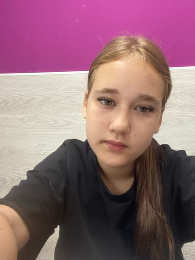 A young woman with light brown hair pulled into a ponytail is looking directly at the camera. She is wearing a black t-shirt. She has green eyes and is wearing false eyelashes. The background consists of a bright pink wall and a light wood-paneled wall. There is a small, circular, patterned object on the wood-paneled wall to the left of the woman. The lighting appears to be from overhead. The image is a selfie taken at eye level. The woman is positioned in the center of the frame. The overall impression is casual and personal. There are no discernible elements that would indicate the specific location in the city of Vysoki, Russia.