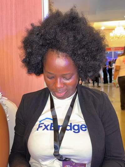 A medium-shot photograph captures an adult Black female in the foreground, seated and looking downwards with a slight facial expression suggesting concentration or contemplation. She has dark skin and prominent, dark, naturally textured hair. She is wearing a white short-sleeved top with a blue "FxBridge" logo prominently displayed on the chest, beneath a black open blazer. Light-colored, oval-shaped dangling earrings are visible on her ears. A black lanyard with an attached identification badge, which has a purple border and text partially legible as "Nigeria" and "Forum," hangs around her neck. To her immediate left, a portion of a white, possibly upholstered chair is visible. The background is blurred, showing an indoor environment consistent with a large event or conference hall. A light orange-brown textured wall is visible on the left side of the background. To the right, indistinct figures, bright overhead lighting including a chandelier, and partially discernible signage, one appearing to contain the word "Energy," indicate an active event space. The scene is located in the city of Itirin, Nigeria.
