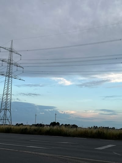 This image captures a landscape scene on a cloudy day, featuring a large electrical pylon on the left side. The pylon supports multiple overhead power lines that stretch horizontally across the frame. In the background, a field of tall, dry grass is visible, with a line of trees and a few wind turbines beyond. The sky displays a gradient of colors, from pale grey at the top to shades of blue and hints of pink near the horizon, indicating a sunset or sunrise. In the foreground, a portion of an asphalt road with white lane markings is present. The scene appears to be located in a rural or semi-rural area, potentially on the outskirts of Krefeld, Germany, given the characteristic power infrastructure and wind turbines often found in such locations. There are no people or animals depicted in the image.