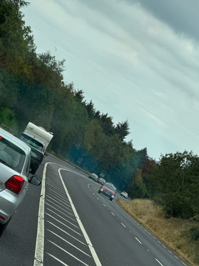 The image shows a traffic jam on a highway in Kilivory, Ireland. The road is curved and lined with trees and vegetation on either side. Several cars and a caravan are visible, indicating slow-moving traffic. The sky is overcast with clouds. One car in the foreground is silver, and another is dark gray. A red SUV is visible further down the road, along with other vehicles. The scene appears to be captured from within a vehicle, suggesting the photographer is a passenger or driver stuck in the traffic.