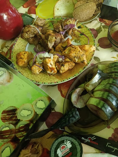 The image shows a table laden with food, possibly a picnic or a gathering at a residence in Pivdenne, Ukraine. The spread includes several dishes: a plate of grilled meat skewers with onions and spices, a plate of sliced smoked mackerel, and a box of sushi rolls. In the background, there are slices of bread and a red pitcher. The table is covered with a floral-patterned tablecloth. There are no people visible in the image, nor are there any specific indicators of the exact location within Pivdenne beyond the presumption of a domestic or outdoor dining setting. The lighting appears to be natural, suggesting daylight.