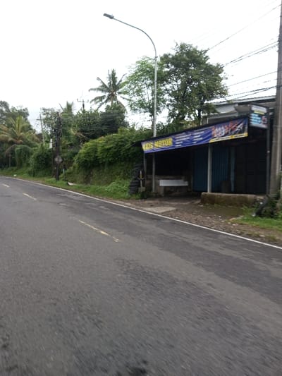 The image presents a roadside view in the city of Vaatharawattai, Sri Lanka, looking forward along a two-lane asphalt road under an overcast sky. The road, featuring faint yellow dashed lane markings, occupies the foreground and midground, sloping slightly upwards from left to right. To the left of the road, a steep, heavily vegetated embankment rises, featuring dense green foliage, several palm trees, and a utility pole with multiple wires.
On the right side of the road, a narrow gravel shoulder precedes a concrete slab leading to a single-story commercial structure. This building has a dark, open front section with a blue corrugated metal back wall visible inside. A prominent blue banner is affixed horizontally across the front facade, displaying 'AZIS MOTOR' in yellow capital letters. Below this, smaller white text, identified as Indonesian/Malay, includes words such as 'Tersedia' (Available) and 'Kaca Film' (Window Film), indicating a potential automotive or film-related service. To the left of the shop's entrance, a stack of worn vehicle tires is visible on the ground, adjacent to a white concrete bench or counter-like structure. A dark utility pole with wires stands immediately to the right of the building, and a tall, slender street light pole with a curved arm extends over the road from the right side of the frame, positioned near the commercial structure. No individuals or active vehicles are present in the scene.