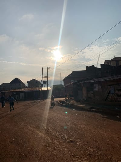 The image depicts a street scene in Enugu, Nigeria, characterized by a dusty, unpaved road under a bright sky with visible sunbeams and scattered clouds. Several buildings, some appearing residential and others commercial with corrugated metal roofs, line the street. Power lines and a transformer are prominent on utility poles.
In the foreground, two individuals are walking or running on the road, their figures silhouetted against the light. A third person is also visible further down the road. The environment appears to be a bustling area with shops and possibly a market, indicated by the structures and the presence of people. The overall impression is a snapshot of daily life in a developing urban setting.