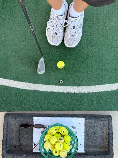 An overhead shot shows a person's feet wearing white New Balance running shoes on a green artificial turf golf practice area. A golf club, identified by the text "RCH 65", is positioned to the left of the frame, with its head near the turf. Two yellow golf balls are visible on the turf, one closer to the club and the other further away. A white line curves across the turf in the mid-ground. In the foreground, a dark mat contains a pair of sunglasses and a green plastic basket filled with yellow golf balls. A white sign with text is also present on the mat. The location is a golf driving range.