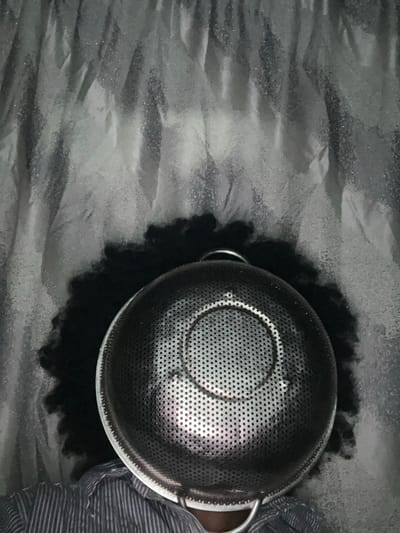 The image depicts an individual in Iranla, Nigeria, whose face is completely obscured by a large, circular, perforated metallic colander. The colander, appearing to be stainless steel, features numerous small, uniformly distributed holes and a concentric ring pattern on its interior base. Two metallic wire handles are visible on opposite sides of the colander's rim, with one partially visible at the bottom. Dark, curly, voluminous hair is visible around the upper and side edges of the colander. The individual is wearing a light-colored shirt with horizontal stripes, visible on the lower left. The background consists of a textured, light grey material, likely fabric or a curtain, displaying vertical folds and a subtle reflective sheen.