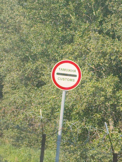 A circular sign with a red border is mounted on a pole in front of a dense green leafy background. The sign has white text on a white background. The top line of text is in Cyrillic, and the bottom line is in English. Both lines read "CUSTOMS". A thick black horizontal bar separates the two lines of text. Barbed wire fences are visible in the foreground and background, indicating a border crossing or restricted area. The overall scene suggests a border checkpoint in Ilek, Russia, a city located near the Kazakh border. The lighting is bright, suggesting daytime.
