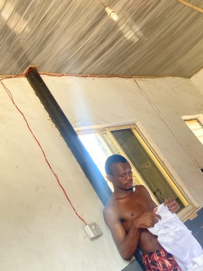 The scene depicts an interior room in Agege, Nigeria, illuminated by natural light from two windows located on the background wall. A single individual, a shirtless male with dark hair and stubble, is present in the center-right of the frame. He wears patterned shorts (red, brown, black, and white) and is focused on handling a crumpled white fabric or plastic object with both hands.

The room features light-colored, textured walls, possibly plaster or concrete. A dark, possibly black, pipe or conduit is angled diagonally upwards from the lower-left towards the ceiling, where it passes through a gap in the wall. Exposed red electrical wiring is secured along the top perimeter of the walls with white clips, and a white electrical outlet is mounted on the left wall. A single, unlit, bare light bulb hangs from a beige cord attached to the light-colored, horizontally paneled ceiling. Bright sunlight streams through the windows, casting strong highlights and creating visible lens flare.