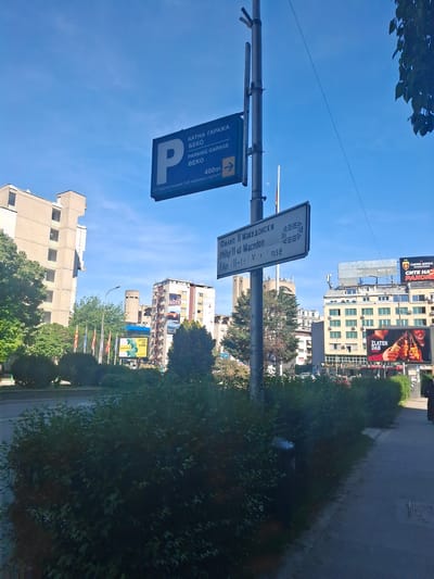 The image shows a street scene in Skopje, North Macedonia, featuring street signs. A parking sign indicates "Parking Garage BEKO, 400m." Another sign references "Philip II of Macedon." Buildings, including those with billboard advertisements, are visible. Trees and greenery line the street.