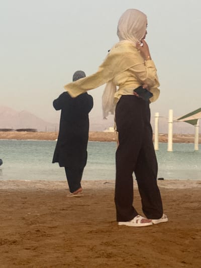 The image captures two individuals by a body of water, possibly the sea, with a sandy shore in the foreground. The scene is set outdoors, with a bright, overcast sky suggesting daytime. In the background, there are faint outlines of hills or mountains. A structure with white horizontal bars and a green and white striped element is visible near the water's edge.
The primary subject is a person wearing a light yellow long-sleeved top and dark trousers, with a light-colored hijab. This individual is holding a dark object, possibly a phone, in their right hand and has their left hand near their neck. Next to them, lower in the frame, is another person dressed in a black abaya or similar garment, with their back to the camera.
The orientation of the image is unusual, with the subjects appearing sideways. The person in yellow appears to be standing or walking on the sandy area, while the person in black is also on the sand, possibly bending or sitting. The activity appears to be casual, perhaps a stroll or a moment of rest by the water.
There is no visible text in the image. The lighting suggests it is daytime, but the overcast conditions prevent distinct shadows or a specific time of day from being identified. The overall mood is calm and relaxed, as people enjoy the coastal environment. The location context provided points to Tamar Regional Council, Israel, which is consistent with a coastal setting.