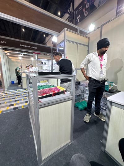This image captures an indoor exhibition hall scene within an assumed trade show environment in Jaipur Municipal Corporation, India. The setting features multiple booths constructed with aluminum frames and light wood-grain panels, defined by dark grey carpeted flooring.
In the foreground right, a male subject stands, facing slightly left with hands on his hips. He wears a white collared shirt, black pants, grey sneakers, and a black beanie, with a pink lanyard and ID badge visible around his neck. To his left, in the midground, another male subject stands behind a glass-topped display case, facing left and away from the camera. He wears a white vest over a light-colored long-sleeve shirt and dark pants.
The central display case, constructed of aluminum and glass, showcases multiple trays filled with colorful beaded strands or small gemstones, predominantly green, pink, and red. A white sign labeled "SURVEY FORM" is partially visible on top of this case. Behind it, another glass display case contains various pieces of jewelry, including necklaces on black display stands.
Further into the background left, within a booth labeled "BANGKOK HOUSE" with booth number "2L27," two male subjects are visible. One, in a green shirt, faces away from the camera, while the other, in a white shirt with a lanyard, leans over a display counter. A patterned section of flooring with grey, black, and yellow stripes leads into this booth.
The background right shows stacked clear plastic storage boxes. An "EXIT" sign with an arrow is visible in the upper right background, alongside a partially visible sign that reads "The Dec...". The ceiling features exposed dark structures and bright overhead artificial lighting. No direct interactions between the subjects are distinctly depicted; they appear to be attending to their respective booths or observing the surroundings.
