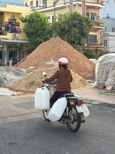 A person wearing a brown hoodie and a white and red helmet rides a Honda motorcycle on a paved road in South Quy Nhon Ward, Vietnam. The motorcycle is loaded with two large white plastic containers, one on each side. The rider is looking forward. In the background, there are piles of sand and gravel, construction materials, and multi-story buildings. The sky is visible in the upper part of the image.