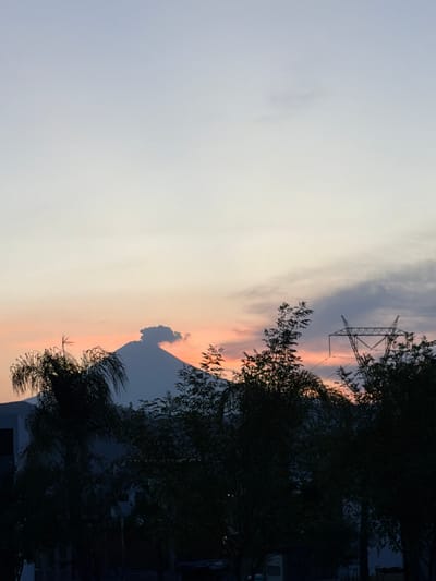 The image shows a view of a sunset in Santa Clara Ocoyucan, Mexico. A large mountain is visible in the center of the frame, silhouetted against the orange and yellow sunset colors. In the foreground, there are trees silhouetted in the right side. In the upper part of the image, there is a grey and white sky and a power line. Some buildings are also silhouetted in the lower-right corner.