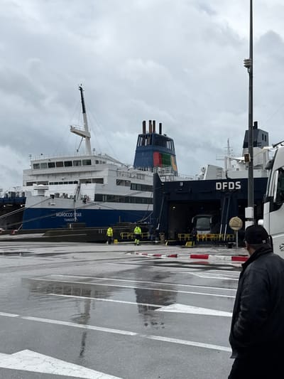 The scene depicts a port terminal in Ksar El Majaz, Morocco, under an overcast sky, with the ground appearing wet from recent precipitation. In the foreground, a male figure with his back to the camera, wearing a dark jacket and a dark cap, stands on wet asphalt. The asphalt features white directional arrow markings and a red-and-white striped concrete barrier.
Two large passenger and vehicle ferries are docked in the midground. The ferry on the left, identified as "MOROCCO STAR" with "TANGER" printed below it, features a blue hull and white multi-deck superstructure. To its right, the "DFDS" ferry is docked, displaying a dark blue hull, white superstructure, and a prominent funnel adorned with horizontal stripes of blue, green, yellow, and red. The "DFDS" ferry's stern vehicle ramp is open, with a white passenger bus partially visible inside the lower deck.
Two individuals wearing yellow high-visibility jackets and dark trousers are present on the dock area between the two ferries, near the "MOROCCO STAR." On the far right, a partial view of a white truck cab is visible, alongside a tall utility pole equipped with an antenna or sensor. Reflections of the ships and sky are visible on the wet paved surface.