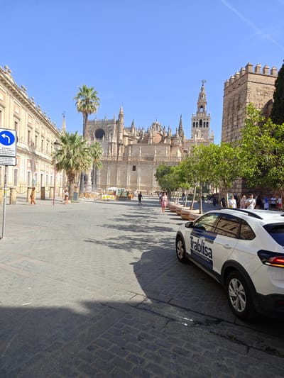 This is a ground-level, outdoor, eye-level shot of a wide, cobblestone plaza in Seville, Spain, with a clear blue sky. In the background, the imposing Seville Cathedral, a UNESCO World Heritage site, dominates the skyline with its intricate Gothic architecture and its iconic Giralda bell tower. To the left, a long, tan-colored building with arched windows and decorative eaves lines the plaza. Palm trees are scattered throughout the scene, adding to the Mediterranean atmosphere.
In the foreground, a white security vehicle with "Trablisa SERVICIOS DE SEGURIDAD" branding is parked on the right side of the frame. Several people are visible walking in the plaza. A blue and white traffic sign, indicating a left turn and restrictions for public service vehicles, is positioned on the left. The ground is paved with dark cobblestones, and shadows from buildings and trees are cast across the plaza, indicating a sunny day.