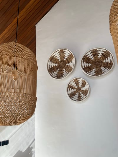 In an interior space, possibly a living room or dining area, located in Kediri, Indonesia, a large, woven rattan pendant light fixture hangs from a sloped, wood-paneled ceiling. The light fixture is intricately woven with vertical rattan strands and features an exposed Edison-style lightbulb. To the right of the light fixture, on a textured white wall, three woven decorative plates with brown and white geometric patterns are mounted. The plates are arranged in a staggered formation, with two larger plates positioned above and to the right of a smaller plate. Shadows from the light fixture cast dappled patterns on the lower portion of the white wall, where a few dark, rectangular tiles are visible near the floor. The overall aesthetic suggests a bohemian or naturalistic decor style.