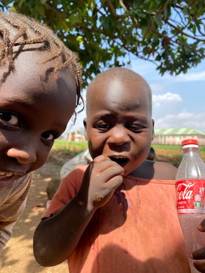 This image, taken in Riyom, Nigeria, captures a vibrant outdoor scene featuring two young children. The photograph is taken from a low angle, looking upwards towards the subjects and the sky.

In the upper right portion of the frame, a child wearing an orange/peach-colored shirt is seen laughing heartily, looking directly at the camera with a wide, open-mouthed smile that reveals their teeth. This child holds a clear plastic Coca-Cola bottle with a red cap. The bottle's label is visible, displaying "Coca-Cola," "TASTE," "SUGAR," "GREAT TASTE," and prominently features the Nigerian flag. The bottle appears to contain a clear, bubbly liquid, suggesting it might be water or a clear soda rather than a traditional cola.

Partially visible in the lower left foreground is another child with braided hair, also looking towards the camera with a gentle smile. This child wears a light brown or tan shirt.

The setting is outdoors under a bright, sunny sky with some white clouds. Lush green tree foliage fills the background on the left side of the image, while a distant, light-colored building with a green roof is visible on the right. Strong sunlight creates distinct shadows, indicating it is likely midday or early afternoon. Both children display cheerful and engaging expressions, conveying a moment of joy and playfulness.