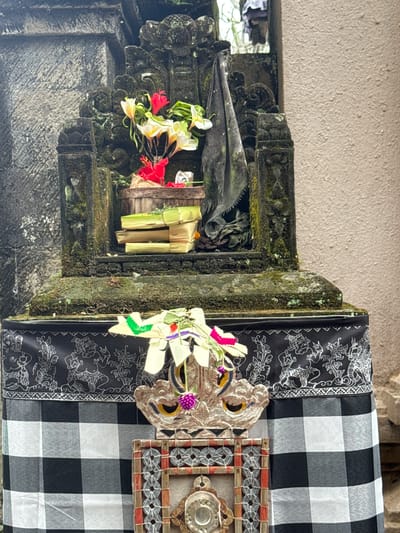 The image shows an outdoor altar in the city of Tegallalang, Indonesia. The altar is made of stone and is covered in moss. It is decorated with flowers and a black and white checkered cloth. There are also some wooden carvings on the altar. The background is a textured wall.