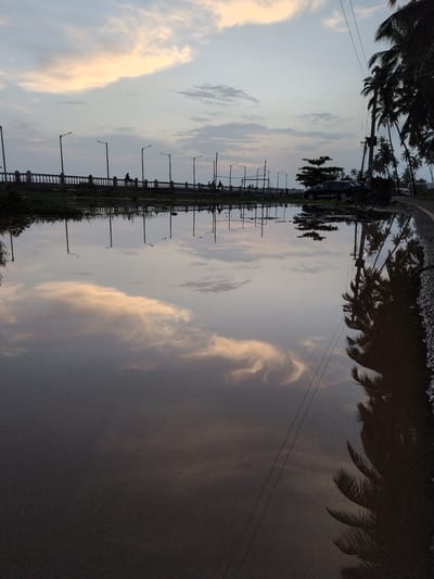 The image captures a reflective scene of a cloudy sky and palm trees in Badem, India. A large puddle or body of water dominates the foreground, mirroring the sky, the clouds tinged with a soft peach hue, and the silhouettes of palm trees lining the right side of the frame. In the background, a pedestrian walkway with ornate railings and lampposts stretches across the image, with several figures walking along it. A dark-colored SUV is parked to the right of the walkway, near some lush greenery and another lamppost. The overall atmosphere is serene, with the setting sun casting a gentle light on the scene.