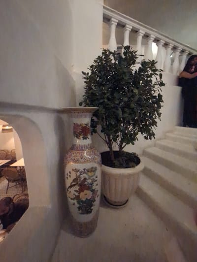 A potted plant and a floral vase are situated on a ledge indoors. In the background is a white staircase. Beneath the ledge is an archway. Through the archway is a glimpse of a tapestry on the wall. It is likely that this scene is taking place in Bengaluru, India.