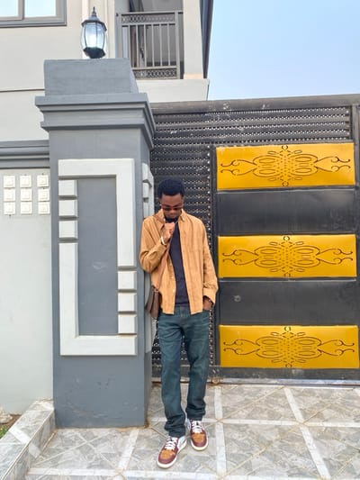 An adult male stands outdoors in the city of Joma, Ghana, positioned to the left of a large black metal gate and against a grey concrete pillar. The man, with short dark hair and wearing sunglasses, is dressed in an open tan long-sleeved shirt over a dark t-shirt, dark blue jeans, and brown, white, and maroon sneakers. A brown patterned cross-body bag hangs over his left shoulder. His left hand is raised to his chin/neck, and his right hand is in his jeans pocket, with his gaze directed downwards.
The grey pillar features white trim and is topped with an ornate dark outdoor lantern. To the right of the man is a prominent black metal gate with two central yellow rectangular panels, each decorated with black scrolled patterns. The gate's upper and lower black sections exhibit a textured or studded appearance. The ground is paved with light grey square tiles arranged in a grid pattern. In the background, a light-colored two-story building with dark window frames and a balcony with a dark railing is visible, under a clear light blue sky.