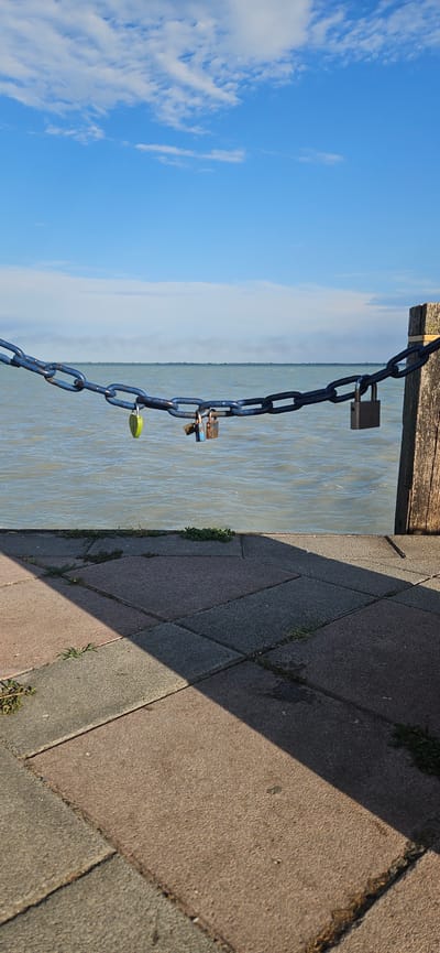 This image captures a waterside scene in Balatongyörök, Hungary. The main subjects are a thick blue chain with several padlocks attached, a body of water, and a paved walkway. The chain hangs from a wooden structure at the top left of the image, extending diagonally downwards towards the water. Three padlocks are visible: one silver padlock is near the top of the chain, another silver padlock is positioned below it, and a small green padlock is near the bottom. The water appears calm with ripples and reflects the bright sky. To the right of the water is a paved area composed of rectangular stones, with a distinct shadow cast across it, suggesting a sunny day. Some sparse green vegetation is growing between the paving stones. The sky is a clear, bright blue with scattered white clouds, indicating fair weather. The overall impression is a peaceful daytime scene, possibly a popular spot for couples to leave "love locks." The time of day appears to be late morning or early afternoon due to the long shadow. There is no visible text in the image.