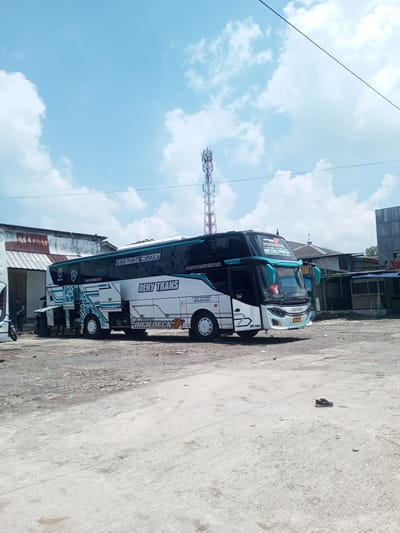 The image depicts a white and black double-decker bus, labeled "Deny Trans" and "Premium Class," parked in a lot. A cell tower rises in the background, under a blue sky with scattered clouds. Several people are visible near the back of the bus, interacting with an open compartment. Buildings with corrugated metal roofs are present behind the bus. A single shoe rests on the concrete lot in the foreground. The location is likely Nawng Hkar, Myanmar, based on the OCR text on the bus.