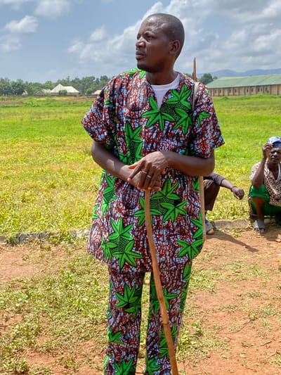 An eye-level, outdoor shot depicts a man holding a wooden stick in Maigizo, Nigeria. The man is facing to the left. He is wearing a shirt with a white under-shirt. The top shirt and matching pants are adorned with green leaf-shaped patterns and a brick-colored textured background. He is wearing a ring on his left hand. Behind him are grasses, land, buildings, and clouds. Behind the man, but slightly out of focus, are two people, one kneeling.