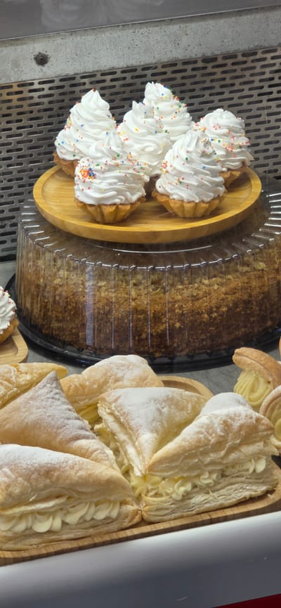 In a bakery display case in Gori, Georgia, several pastries are arranged on wooden trays. In the foreground, triangular puff pastries filled with cream and dusted with powdered sugar are visible. Behind these, a large round cake is covered by a clear plastic dome. On top of the dome, a wooden tray holds small tartlets topped with white cream and colorful sprinkles. The background shows a metal grate, typical of a bakery or food service establishment.