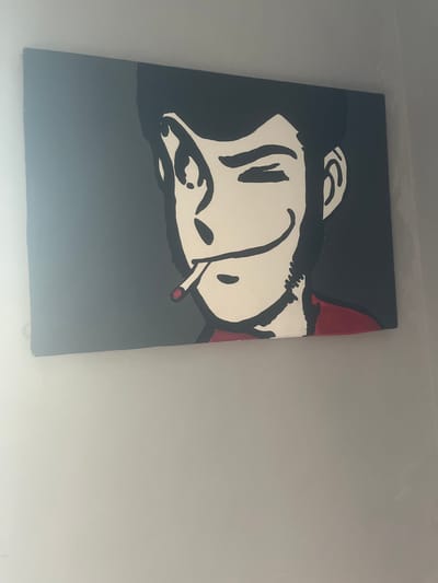 A painting is displayed on a wall. The painting is a stylized depiction of a man's face. The man has dark hair, a red collar, and a cigarette in his mouth. His eyes are closed in a wink, and he has a smirk on his face. The background of the painting is a dark gray. The painting is hung on a light gray wall, and the overall lighting is dim. The image does not contain information about the city of Stornara, Italy.