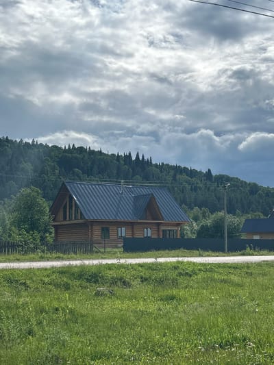 The image depicts a rural landscape with a wooden log house prominently featured in the foreground. The house has a dark, sloped roof and several windows. Behind the house, a dark fence separates it from a lush green forested hill. The sky is overcast with dramatic clouds. In the lower part of the image, a grassy field with scattered wildflowers stretches towards a gravel road. A power line is visible crossing the upper part of the frame. The scene appears to be set in a natural, somewhat remote location. There is no indication of specific people or actions. The location is identified as Usman'gali, Russia.